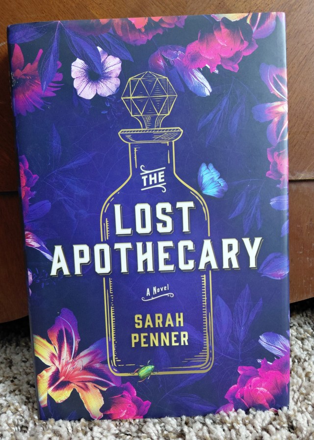 Image is a photo for a book cover for The Lost Apothecary by Sarah Penner. The book cover is a violet color with magenta flowers on it. There's a vial sketched in gold. It looks like a vial that you would store an old perfume or elixir in. 