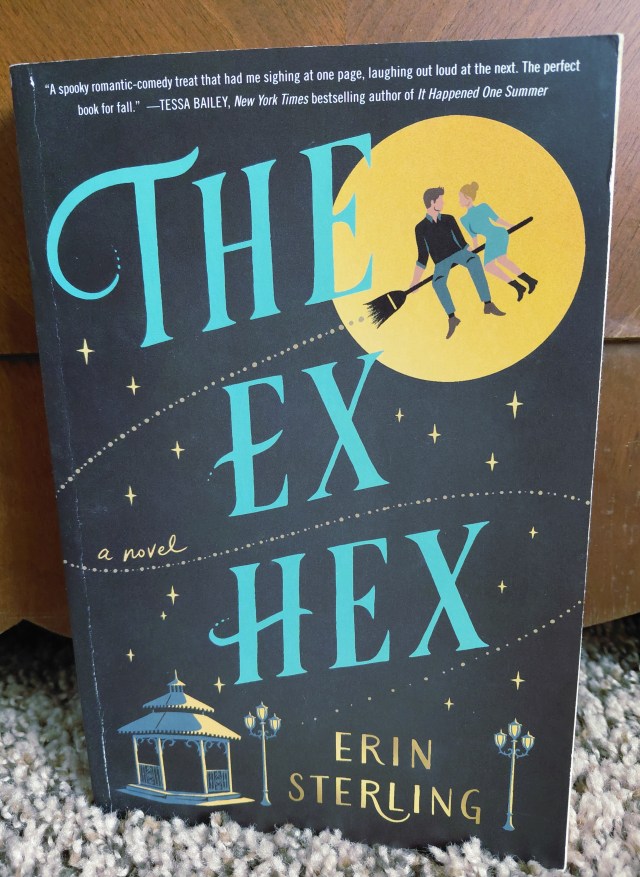 Image is a photo for the book cover, The Ex Hex by Erin Sterling. It shows a yellow moon to the right side of the cover. There's a couple sitting on a broomstick Below to the bottom half of the cover is a Gazebo and a few light fixtures. 