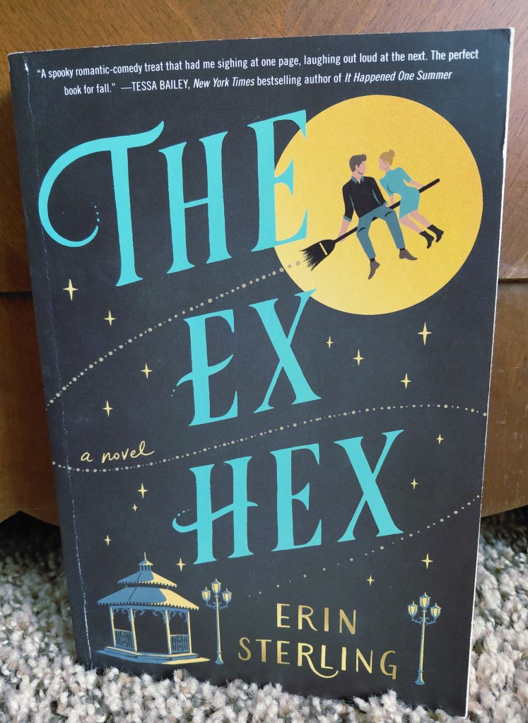 Image is a book cover for The Ex Hex by Erin Sterling. It shows a yellow moon and in front of the moon is a man and a woman sitting on a broom looking at each other. On the bottom of the cover, it shows a gazebo and several lamps. 