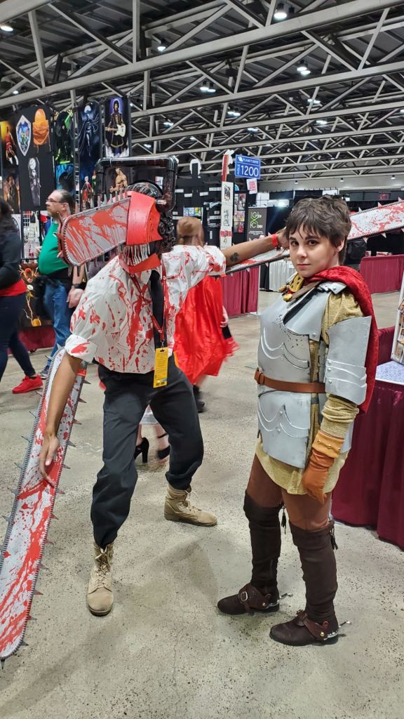 [Image features two cosplayers. The first has a red helmet with a chainsaw protruding from it. There are two chainsaws on both arms. The other cosplayer is in full armor with a red cape]