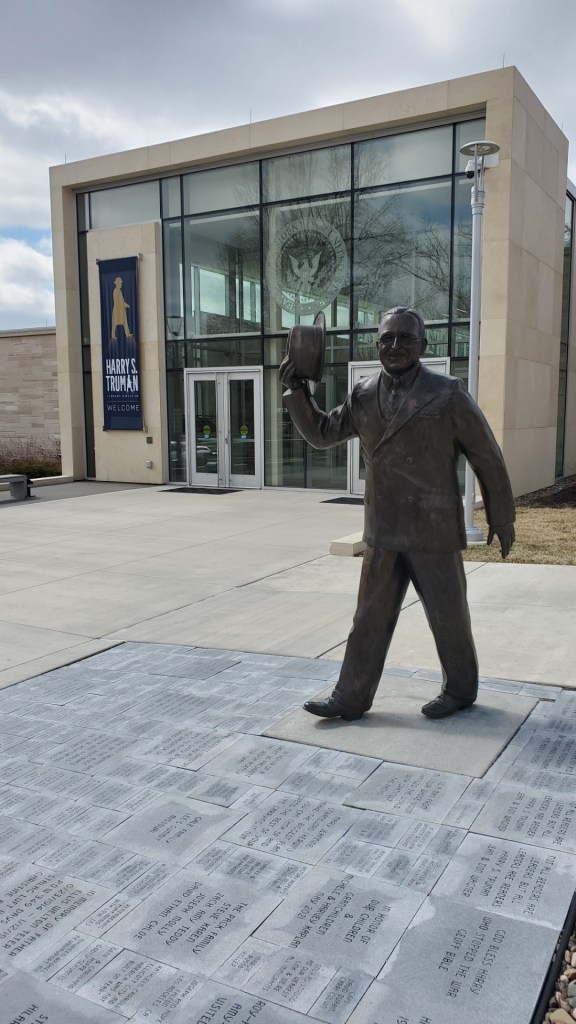 [Image is the front of the Harry. S. Truman Presidential Library. There's a statue of a man smiling with glasses. He's tipping his top hat towards the picture]