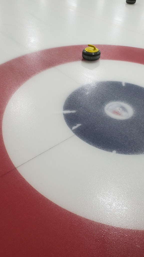 [Image is a ring on ice. There's a red outer ring, a white inner ring, and a black circle. In the red ring, there's a gray stone with a yellow handle on it]