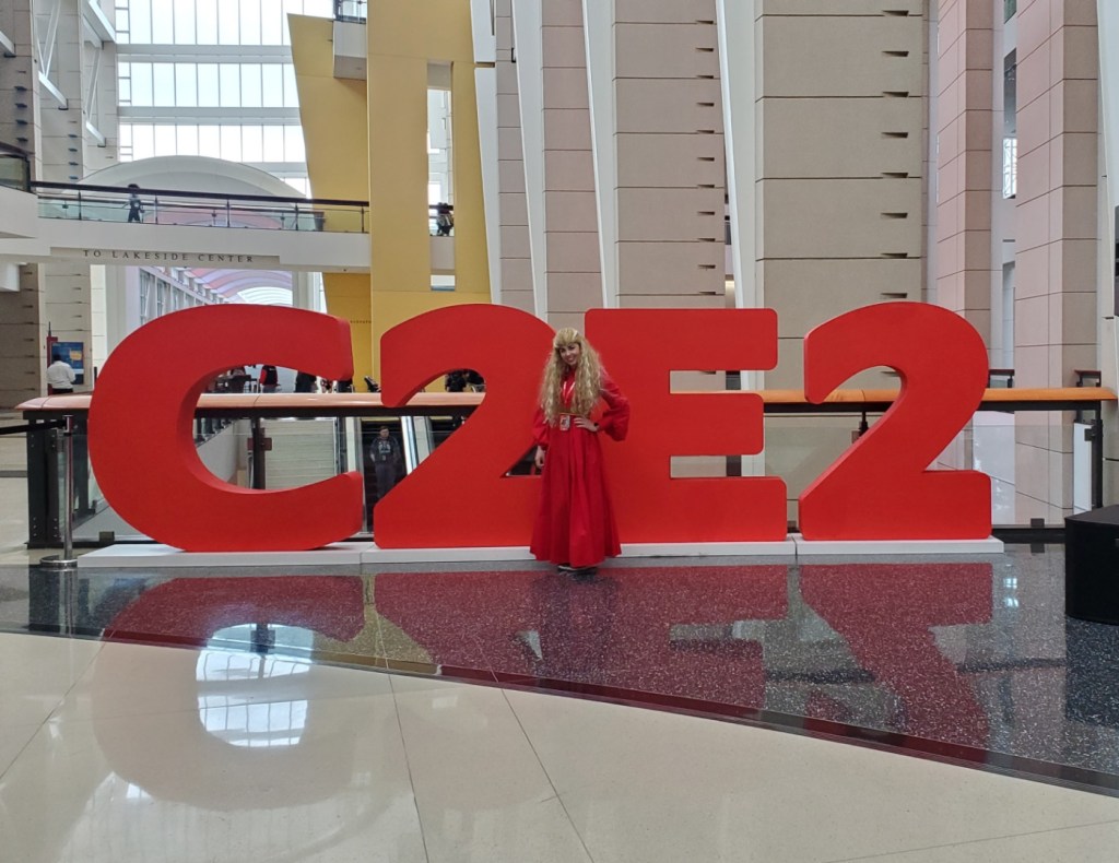 [Image shows a sign that says C2E2. A woman in a red dress, blond wig with her hand on her hip is posing in front of  it]