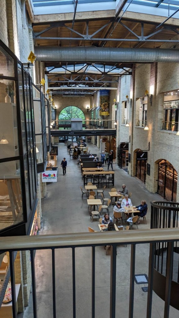 [Image shows a warehouse  type building with several upper walkways. In the middle, are several tables and chairs. There are shops inside with banners pointing out their business] 