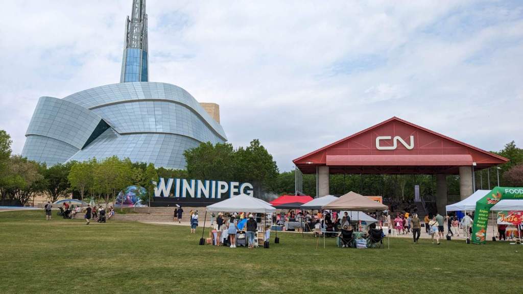 [Image shows downtown Winnipeg. Off to the left, there's a huge blue building with a tower on top. To the right, there's a red stage that says "CN" Several tents are posted below with various vendors selling things]