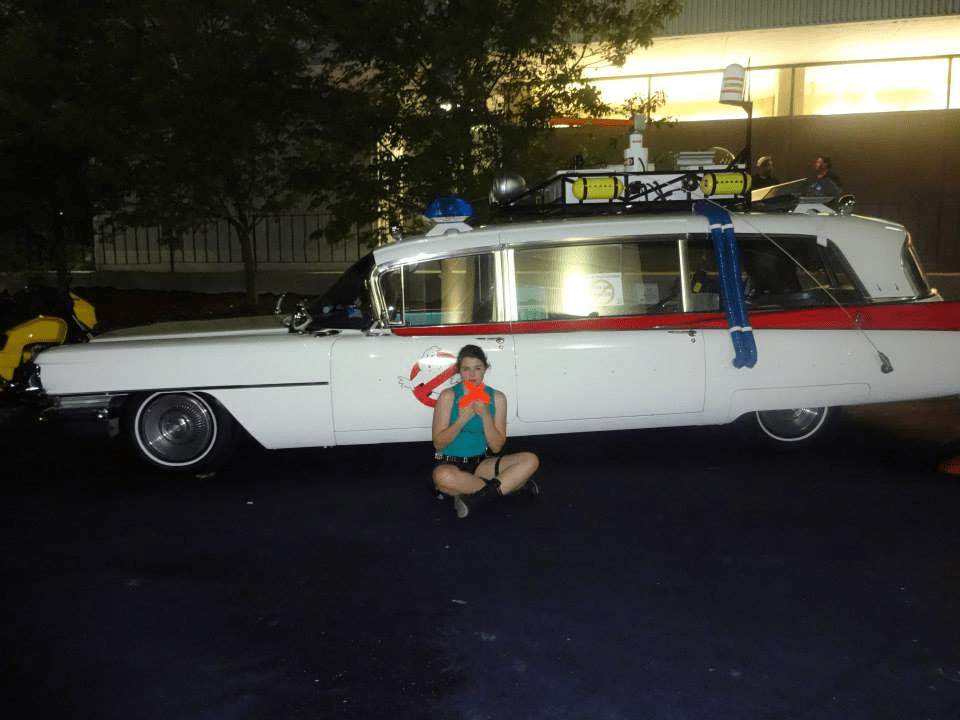 [Image features the ghost busters car. A woman is sitting cross legged in front of the car holding orange plastic guns]