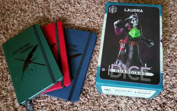 [Image shows a stack of three notebooks. They all say Adventure Journal with a sword and a feather crossed over each other. On the right, it's a box which says Laudna on it. It shows a woman holding black goo and a d20 is sitting on top of it.]
