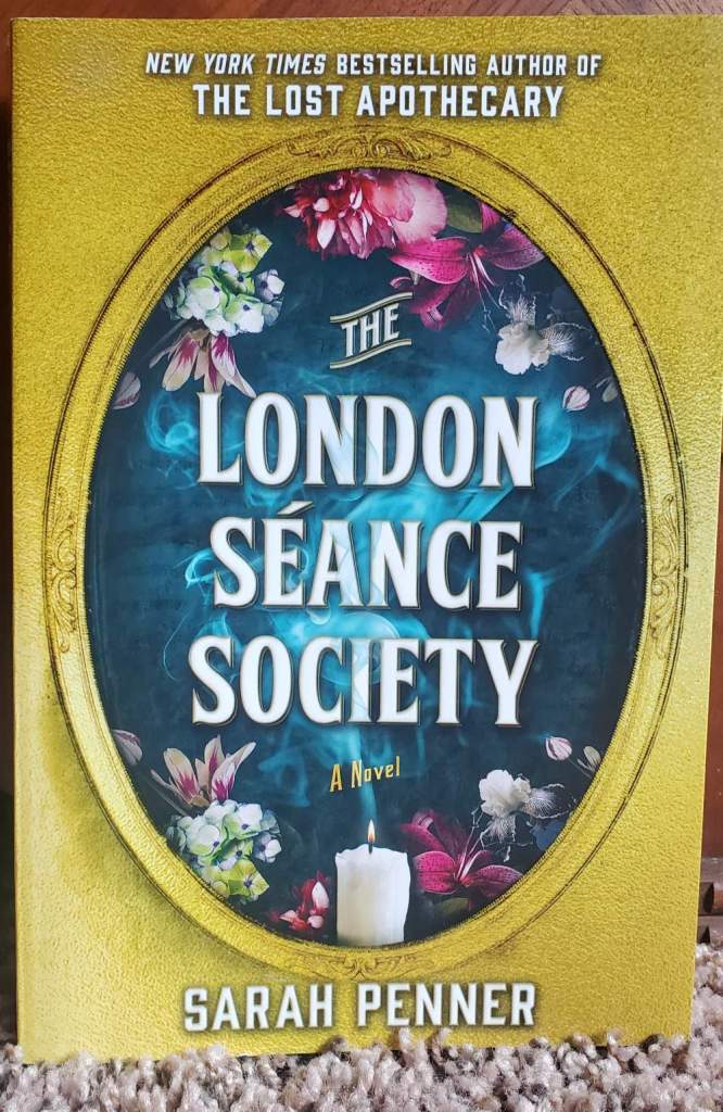 [Image is a book cover. There's an oval shape and in that oval are bright flowers. In the center of the oval is a candle. There are mystical whisps in the air. It says The London Seance Society by Sarah Penner on it]