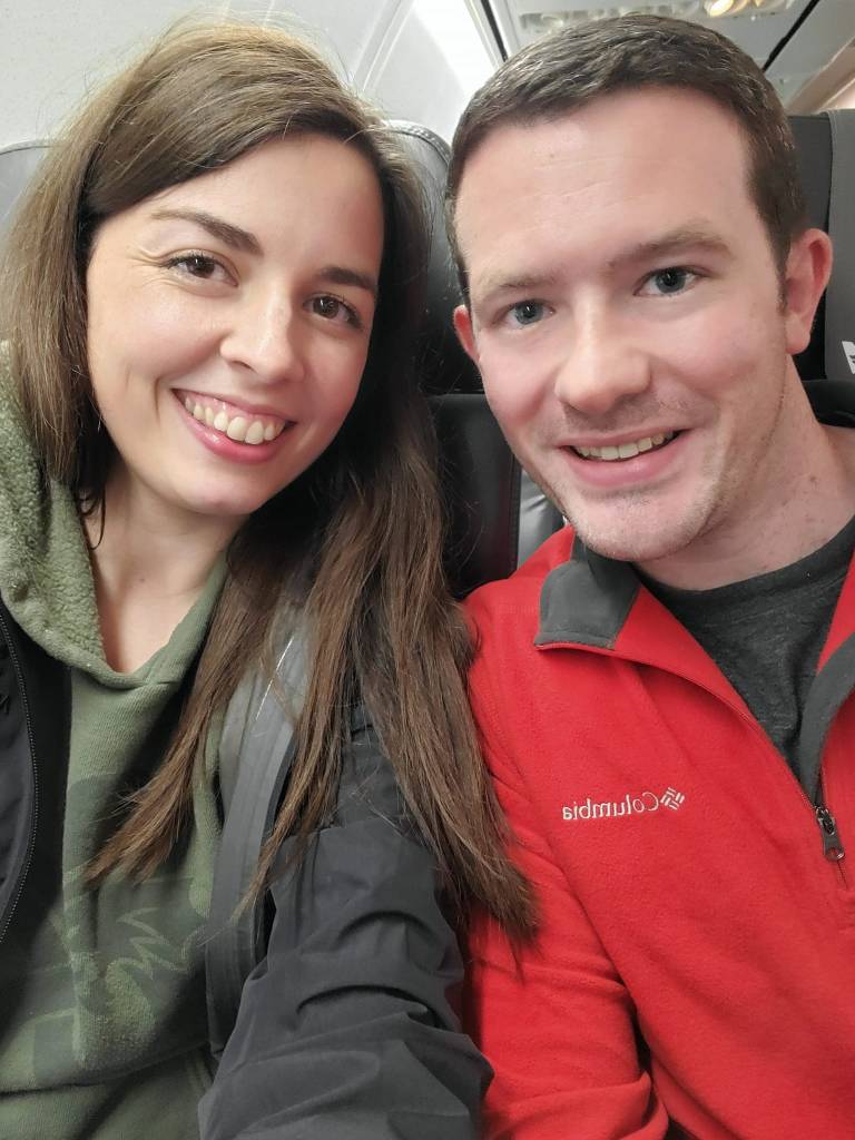 [Image is a selfie of me and my husband. We are both smiling and looking at the camera. We are on an airplane]