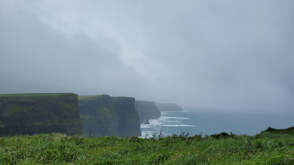 Image is of The Cliffs of Moher. There are several cliffs that are a mix of a bright green and brown. There's water below. It's cloudy, so there is fog on the water where you can't see the end of the water. 