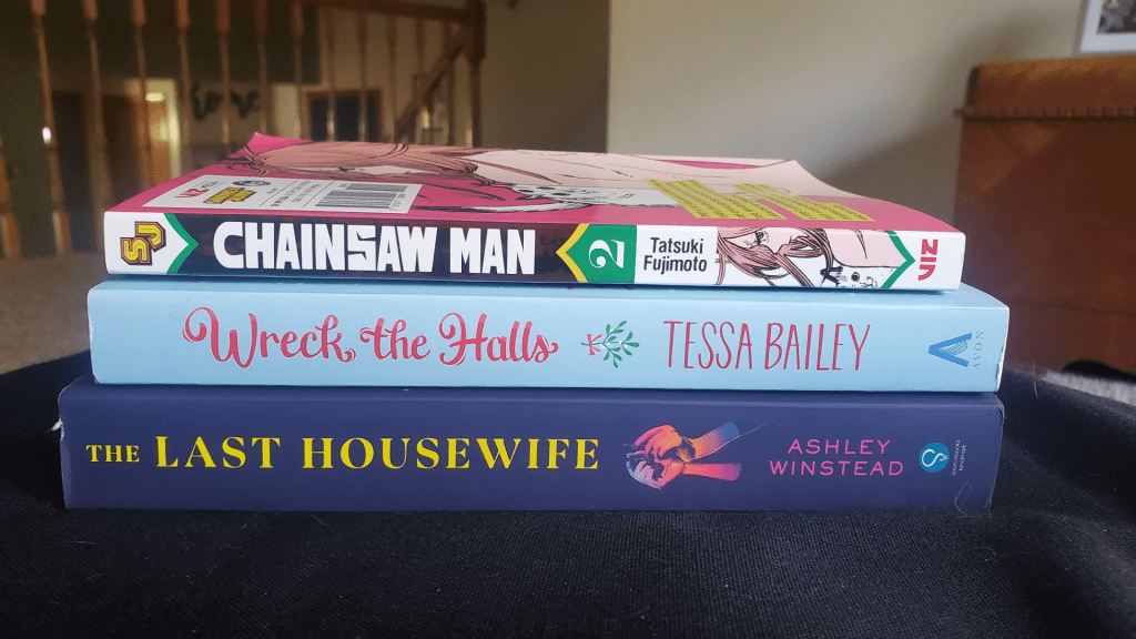 Image shows a stack of three books. The bottom book is The Last Housewife by Ashley Winstead. The middle book is Wreck The Halls by Tessa Bailey and the top book is Chainsaw Man Vol #2.