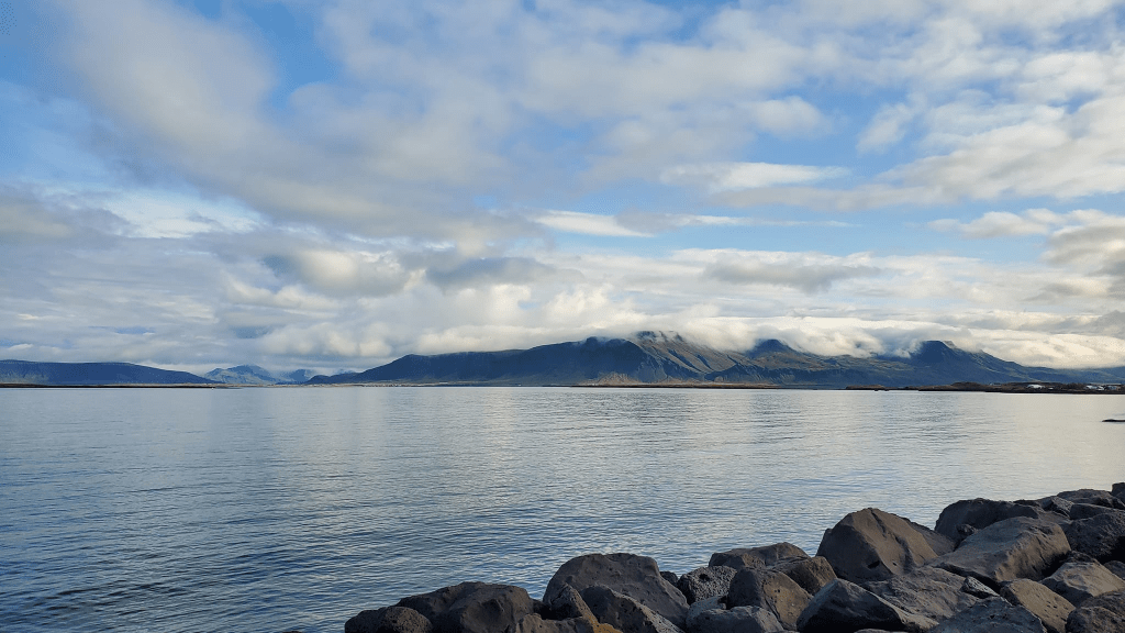 Photo is a distant view of several mountains. The clouds look like they are laying on top of the mountains. There is a body of water that separates the mountains from where we were standing