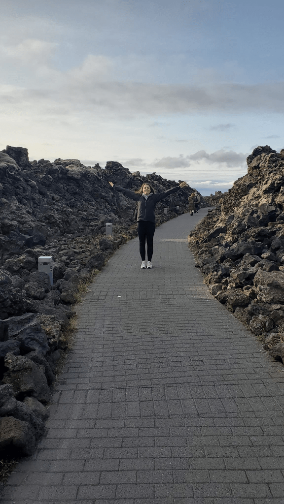Photo is of me standing in the walkway to The Blue Lagoon. There is a gray brick pathway. On either side are these black and white lava rocks.