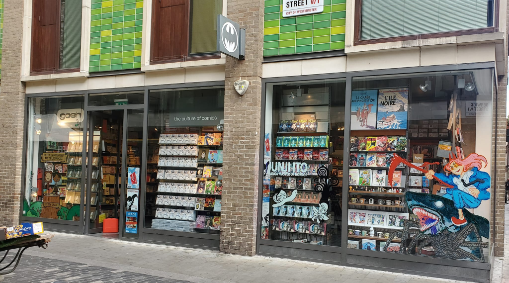 Image is a comic book store. There's a Batman sign that splits the shop windows. There's a sign with gosh in lower case on the left hand window.