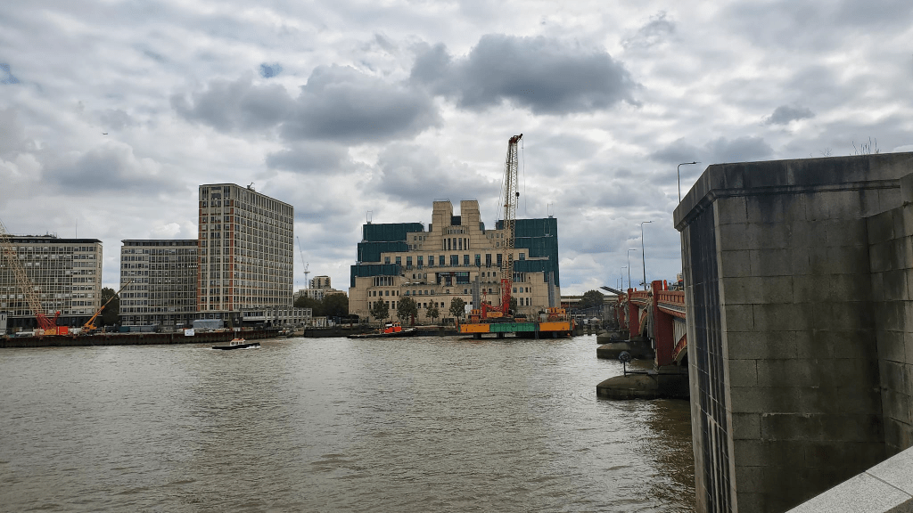 Picture is a building that is across the Thames river. The building has a blue roof. It almost looks like solar panels at the back. It's a brown building with two towers in the back.