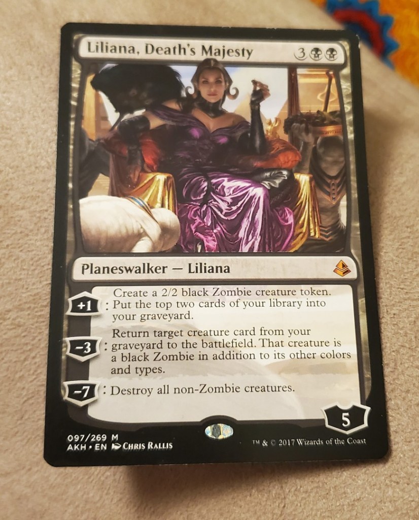 Image is a Magic the Gathering card. It says at the top "Liliana, Death's Majesty" It shows a woman in a purple dress, she has a black fan nearby her. It seems like there are servants nearby her. It says below the art "Planeswalker - Liliana". 