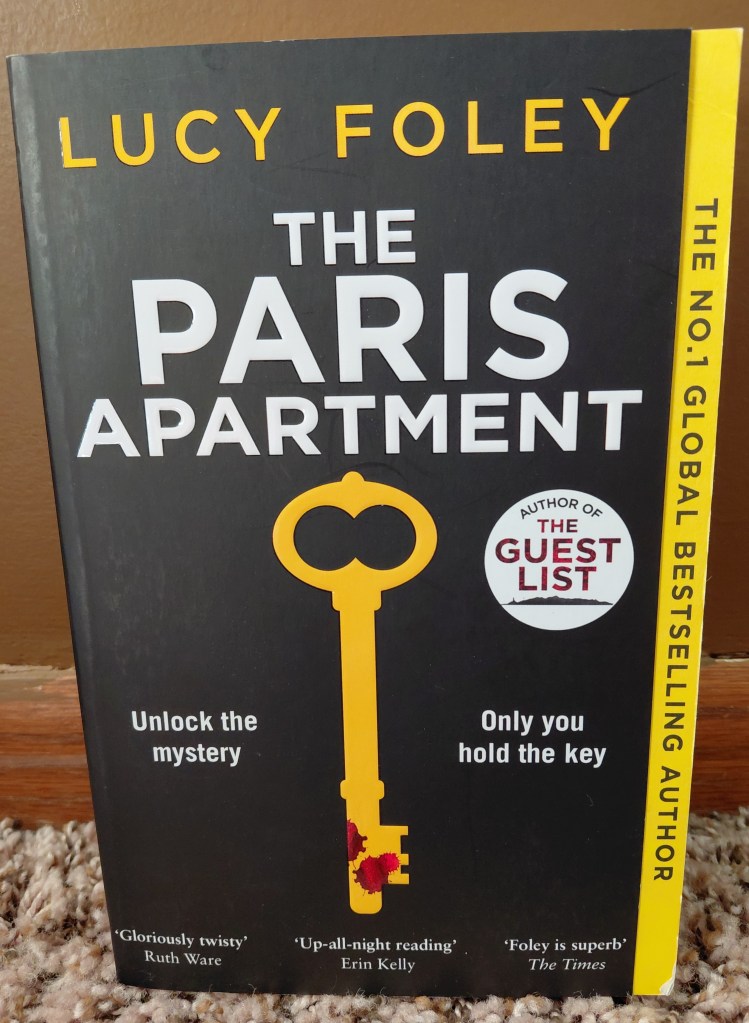 Image is a photo of the book, The Paris Apartment by Lucy Foley. It's a black and yellow cover. There's a yellow key filled in with solid yellow. Towards the bottom of the key are red droplets of blood. By the key it says, "Unlock the mystery" and "Only you hold the key" 