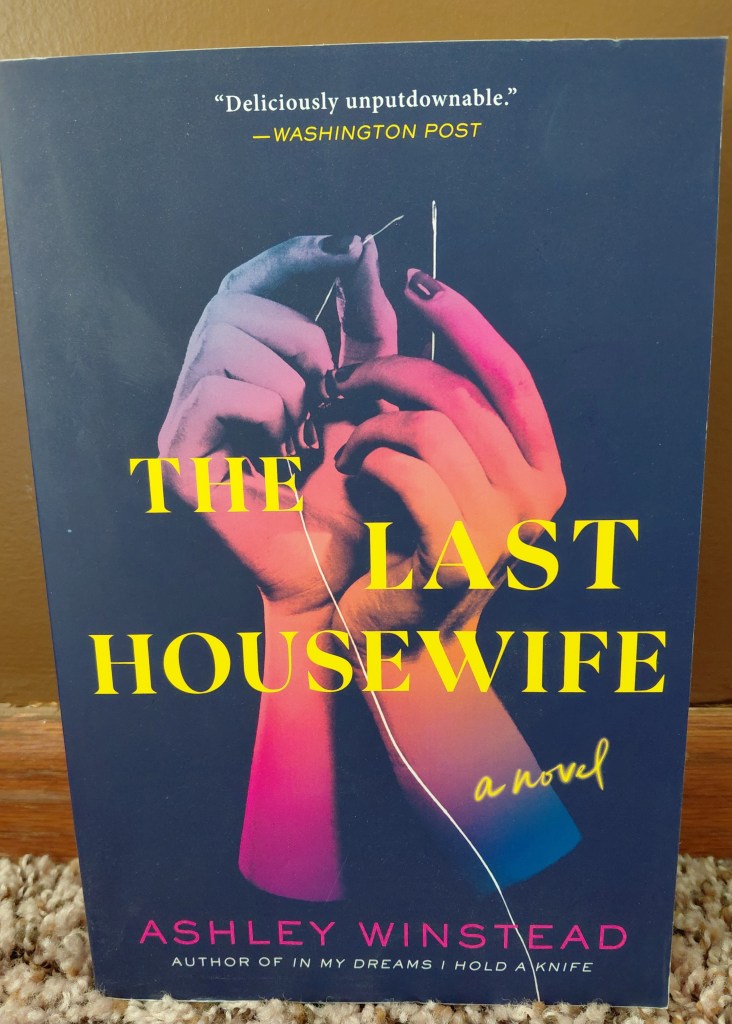 Image is a photo for the book, THe Last Housewife A Novel by Ashley Winstead. The cover is a navy blue and on the front are two hands lit up in purple, pink, and orange lighting. The hands are trying to put a thread into a sewing needle. 