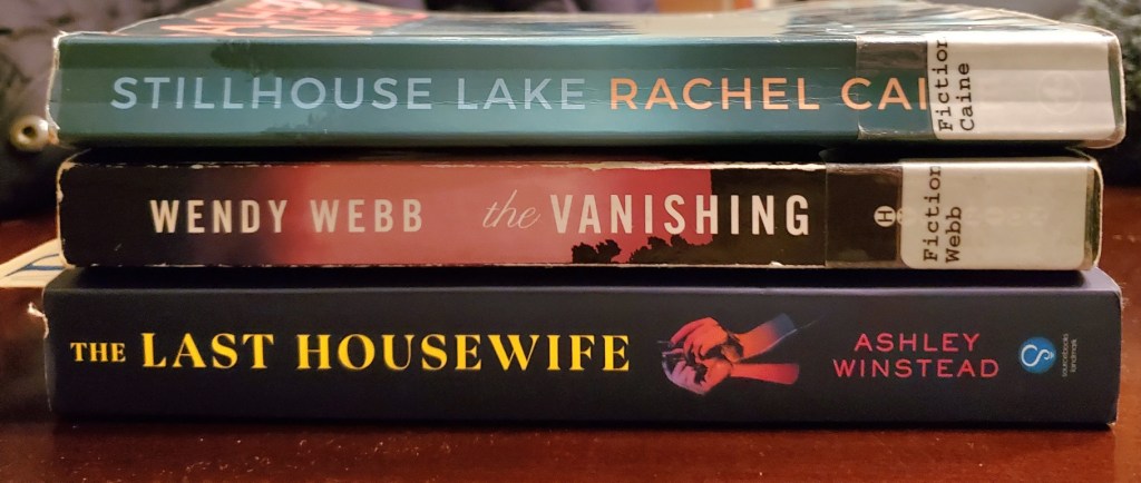 Image is a stack of three books stacked on top of each other. The first book is Stillhouse Lake by Rachel Caine. The second book is The Vanishing by Wendy Webb. The last book is The Last Housewife by Ashley Winstead