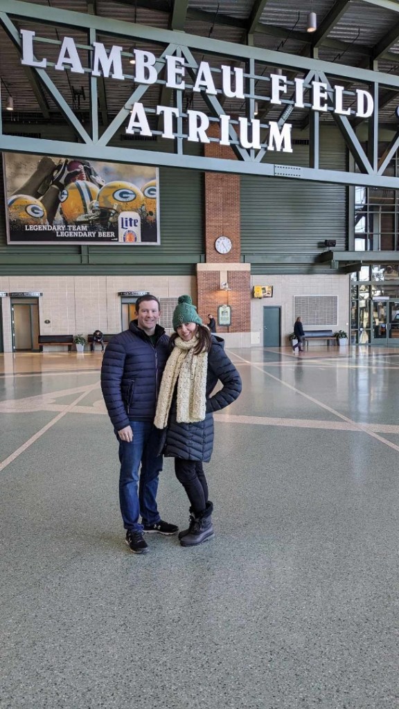 Image is a picture of me and my husband standing inside Lambeau Field. There's the Lambeau Field Atrium above us and we are smiling below the sign. 