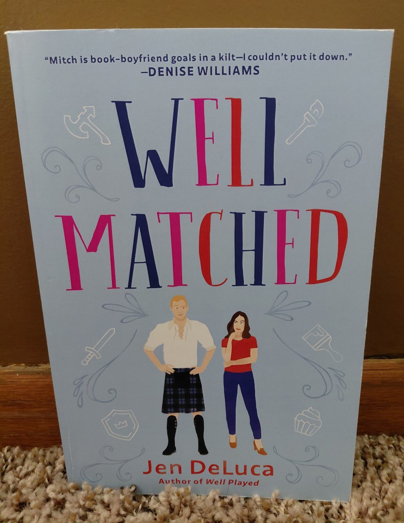 Image is a photo of a book called Well Matched by Jen DeLuca. It's a bright blue cover. There are white symbols etched in almost like a drawn sketch in white. They are an axe, torch, sword, shield, paint brush, and a cupcake. Below the title of the book, there are two people looking side eyed at each other. The man is on the left and he's wearing a kilt and on the right is a woman wearing a red t-shirt and jeans. 