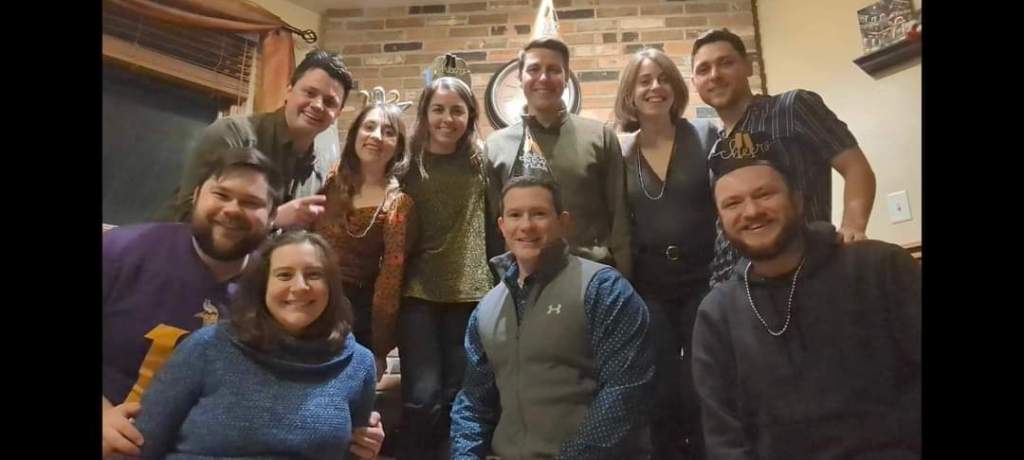 Image is a group friends photo. There are 10 people wearing New Year's accessories. Everyone is smiling