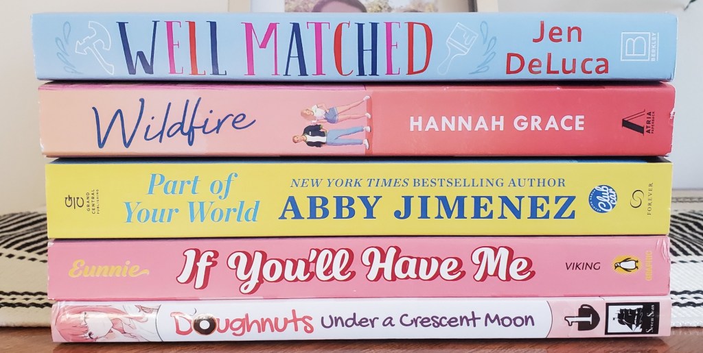 Image is a stack of five books that are laid out by their spine. The first book is Well Matched by Jen DeLuca, the second book is Wildfire by Hannah Grace, the third book is Part of Your World by Abby Jimenez, the fourth book is a graphic novel titled If You'll Have Me, and the final book is Doughnuts Under a Crescent moon