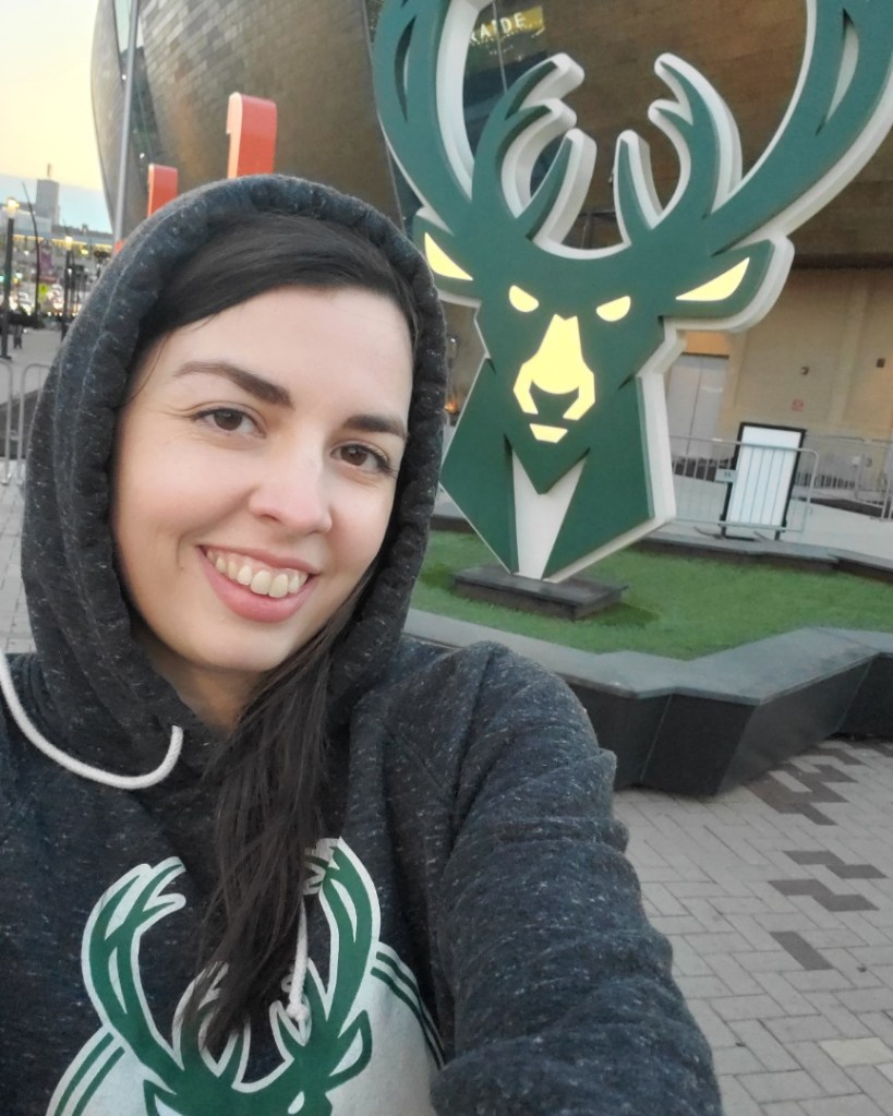 Image is a selfie. I'm smiling at the camera. Behind me, is a glowing statue of a deer with antlers. It's green and yellow. It's the logo of The Bucks. 