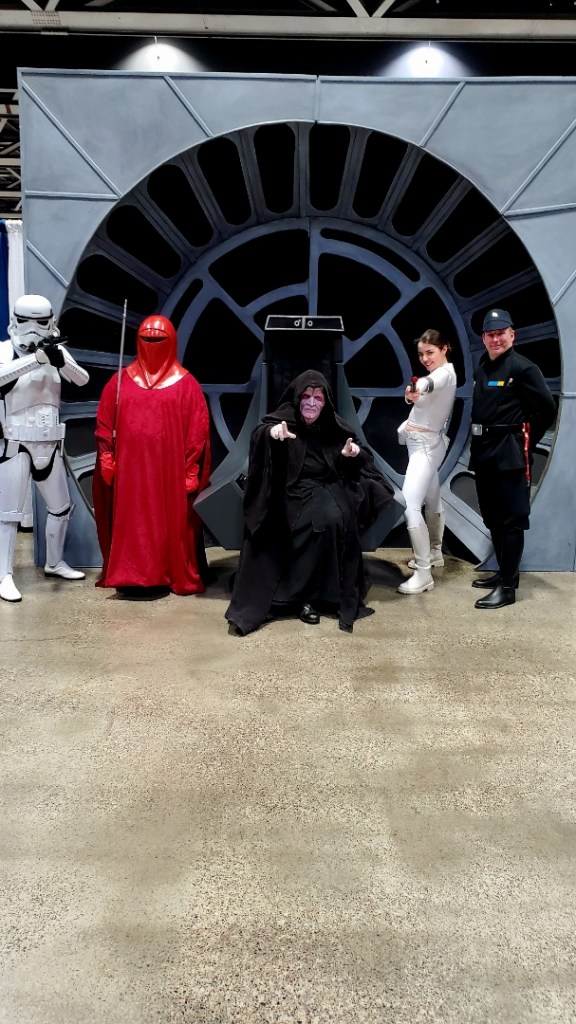 Image is a photo from Planet Comic Con. There are five people in costume. From left to right, there was a Storm Trooper in white armor. There's a red guarrd covered in a red cloak and mask. Emperor Palpatine is in the middle sitting in a throne. I'm next to Palpatine, holding a foam blaster. On the end is an Imperial Officer.