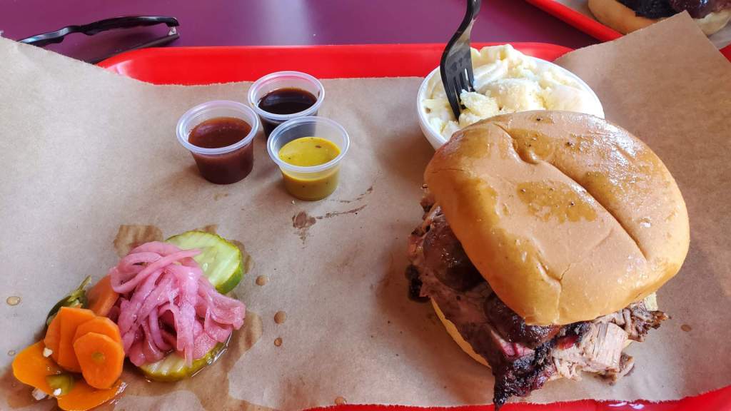 Image is a photo of food. To the right is a BBQ sandwich in a bun. Behind it is a dish of mac and cheese. There are three sauces in three different containers. 