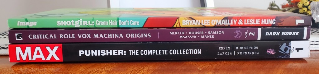 Image is a stack of three graphic novels with their spines showing. The graphic novel list from top to bottom: Snotgirl: Green Hair Don't Care, Critical Role Vox Machina Origins Vol #2, and Punisher: The Complete Collection #1. 