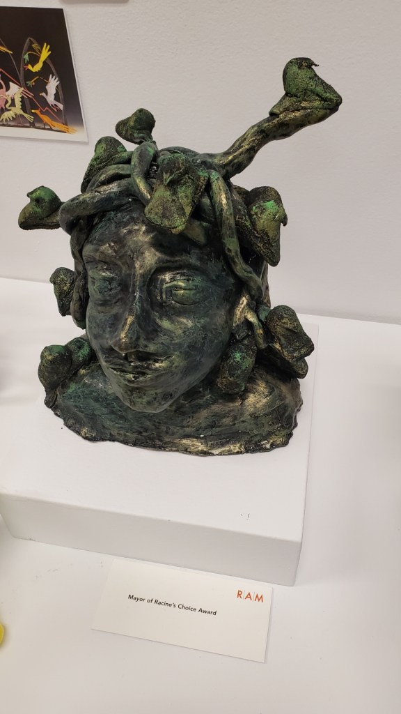 Image is a sculpture of Medusa in a dark black and green. On the ends of her hair are green peeps.