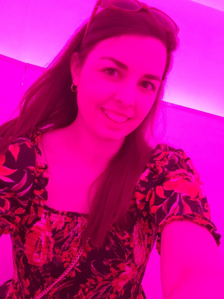 Image is a selfie of me smiling at the camera. I'm under a pink light, so my skin looks pink. I have sunglasses on my head. 