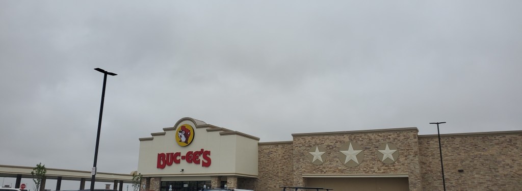 Image is a photo of the Buc-ee's building. It shows three white stars to the right. The logo is a yellow circle with a beaver wearing a red hat. The writing is in red and it says Buc-ee's. 