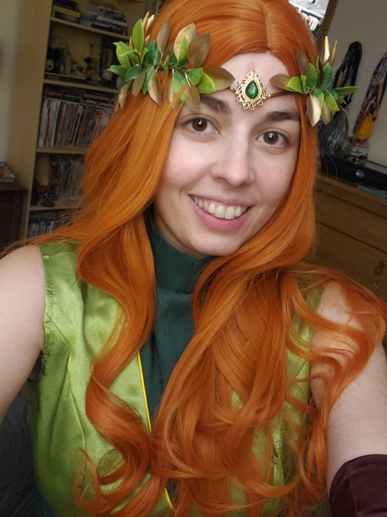 Image is a photo of me cosplaying as Keyleth. I have an orange wig on, green dress, and a floral green crown on. 