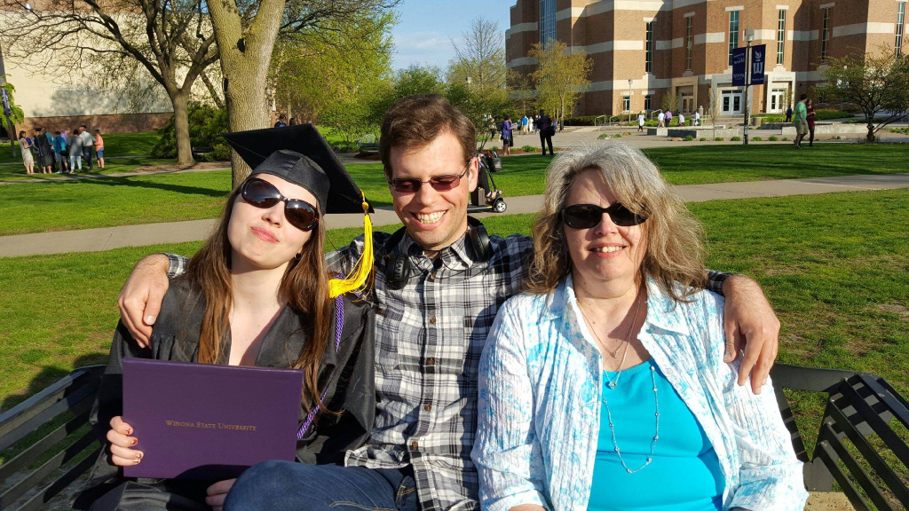 Image is a photo of me, my brother, and my mom. We are sitting on a bench and it is bright outside. We are all wearing sunglasses. I'm holding my purple Winona State University book with my degree. I'm wearing my black cap and gown. 