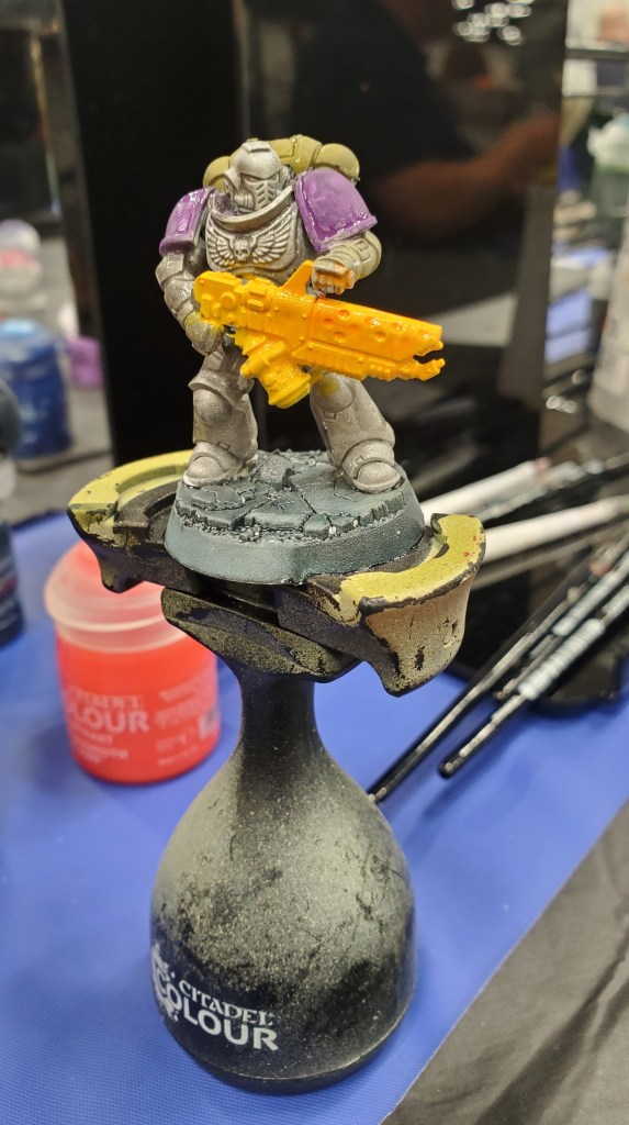 Image is a picutre of a painted Warhammer mini. It's sitting in a pedestal. The shoulder pads are bright purple, the figure is a gray and the figure has a flame thrower that's been painted bright yellow.