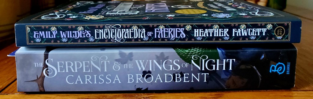 Image is a stack of two books laying vertically on top of each other. The first book is The Serpent & The Wings of Night by Carissa Broadbent. The second book on top of that book is Emily Wilde's Encyclopaedia of Faeries by Heather Fawcett. 