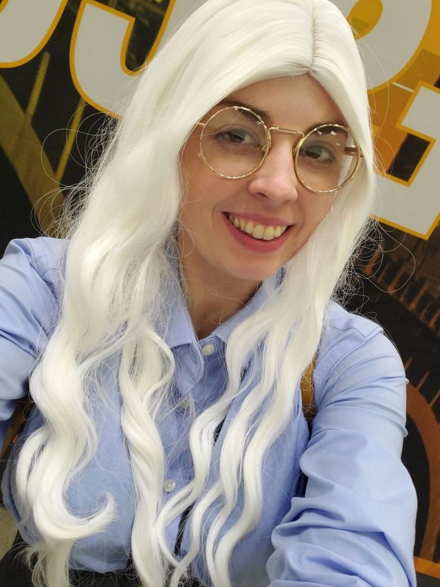 Image is a photo of me smiling at the camera. I'm wearing glasses and a white wig. I'm posing behind a yellow background that reflects Twin Cities Con. 