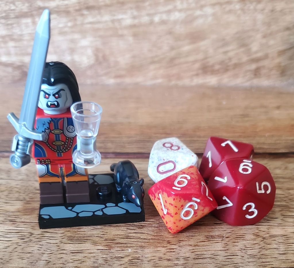 Image is a picture of a LEGO figure. He's holding a sword and a cup. He is clearly a vampire. Next to the LEGO figure are four D10 dice. Two of them are red, one is white and red and the other is red and orange.