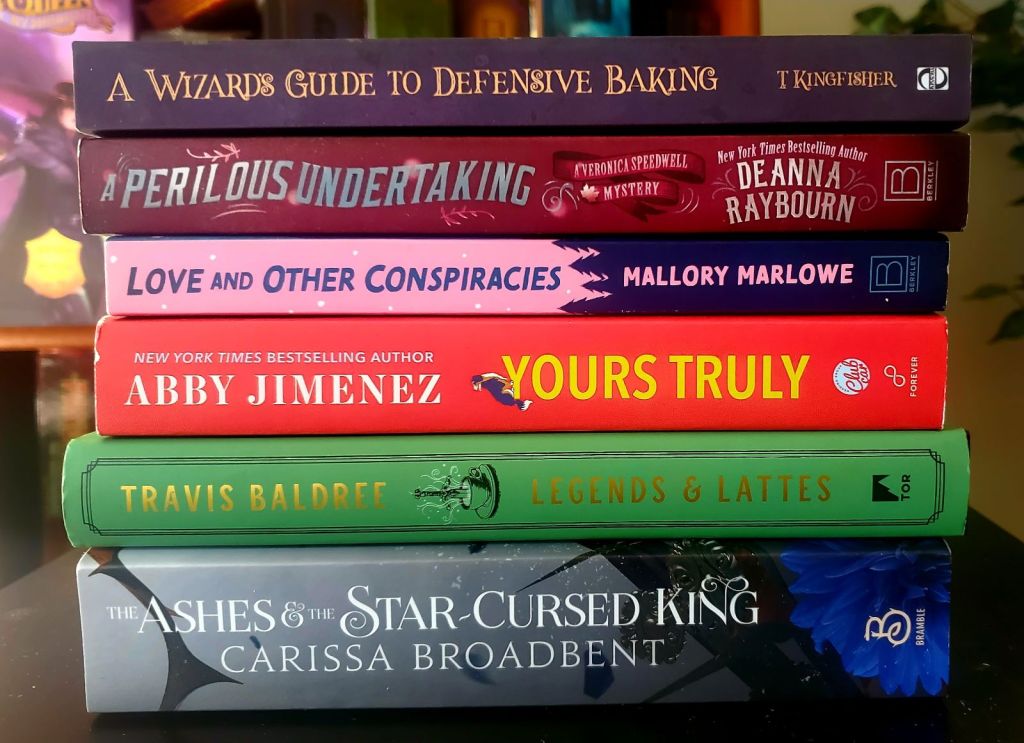 Image is a stack of five books. They are as follows (top to bottom) A Wizard's Guide to Defensive Baking by T Kingfisher, A Perilous Undertaking A Veronica Speedwell Mystery by Deana Raybourn, Love and Other Conspiracies by Mallory Marlowe, Yours Truly by Abby Jimenez, Legends & Lattes by Travis Baldree, and The Ashes & The Star Cursed King by Carissa Broadbent 