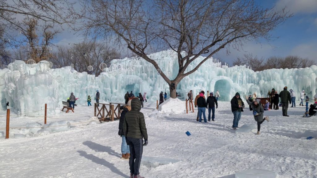 Image is another photo of the ice castle. You can see an opening which leads to a tunnel. There are snow globes hanging from the trees. 