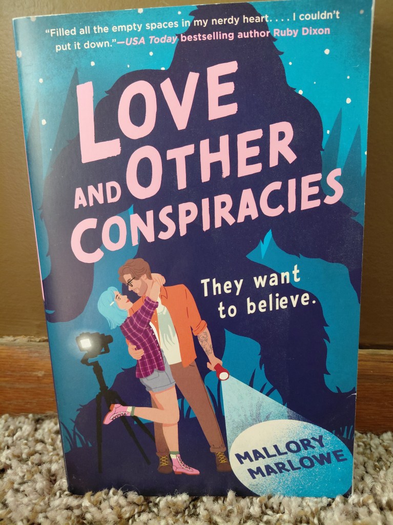 Image is a photo for a book cover for Love And Other Conspiracies by Mallory Marlowe. It shows a couple where the woman has her arms around the man's neck. There's a shadow of Big Foot behind them. 