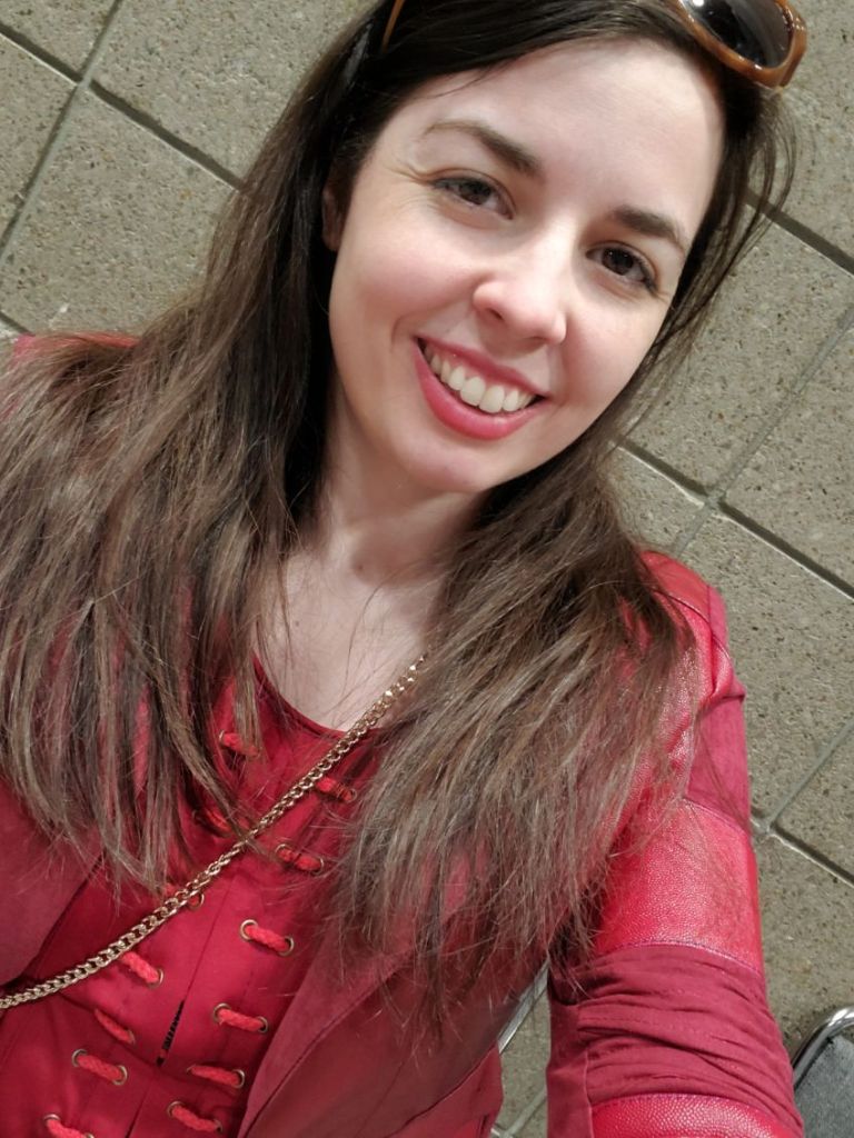 Image is a photo of a woman smiling at the camera. She's wearing all red - red jacket, red top. Her brown hair is long and she has lip stick on.