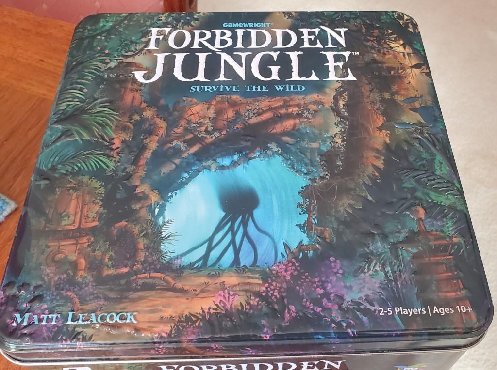Image is a photo of a board game sitting on a table. The board game is Forbidden Jungle. It shows a jungle and a blue tunnel at the center. The shadow within the blue tunnel shows an alien with a black body and several limbs holding it together. 