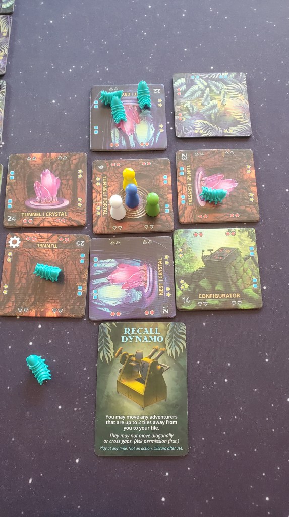 Image shows a group of tiles. There are four tiles with glowing pink crystals on it. In the center, there's a tile with four game pieces on it. There's a card at the bottom that says "Recall Dynamo". The card reads - You may move any adventurers that are up to 2 tiles away from you to your tile. 