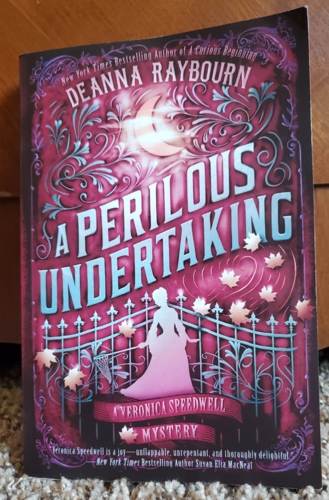 Image is a book cover for A Perilous Undertaking by Deanna Raybourn. This cover shows a silver gate with a grey shadow of a young woman holding a butterfly net. 