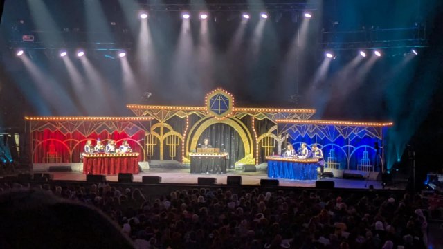 Image is a photo of the stage. There are at least 13 different yellow strobe lights pointing at the stage. There is one half of the stage with a red curtain background and the other half of the stage with a blue certain background. In the middle is a gold, carnival entrance of sorts. At the top if the Critical Role logo. 