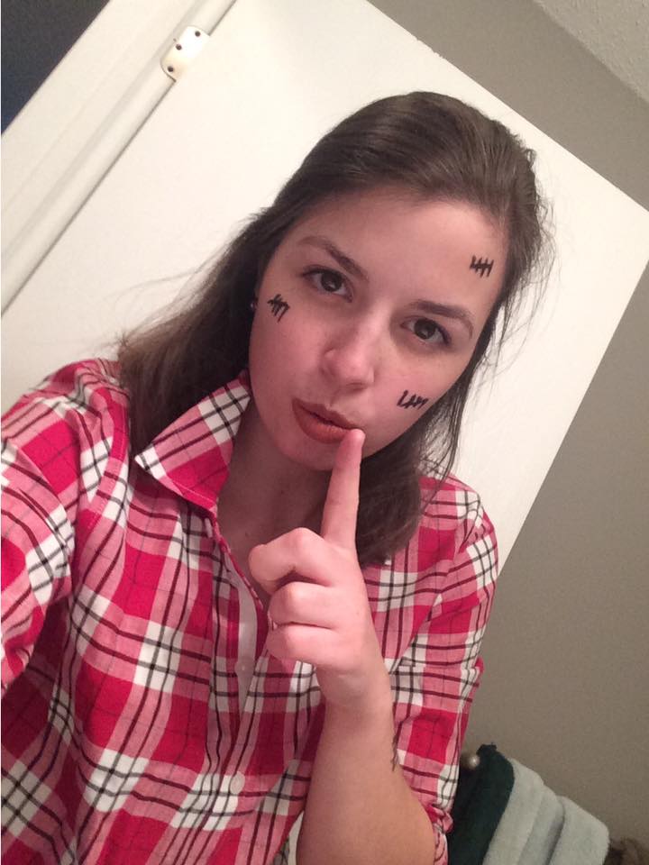 Image is a photo of me taking a selfie. I'm shushing with my finger. I'm wearing a red plaid shirt with black tallies on my face.