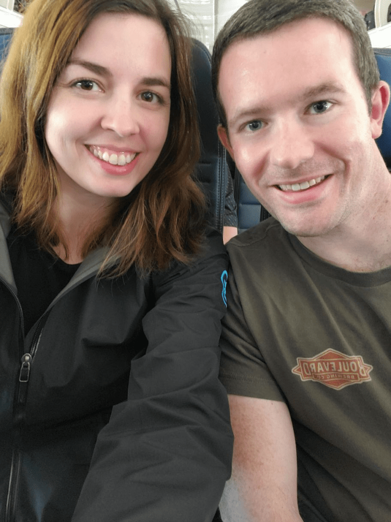 Image is a photo of me and my husband smiling at the camera. We are sitting in our airline seats waiting for our flight to take off. 
